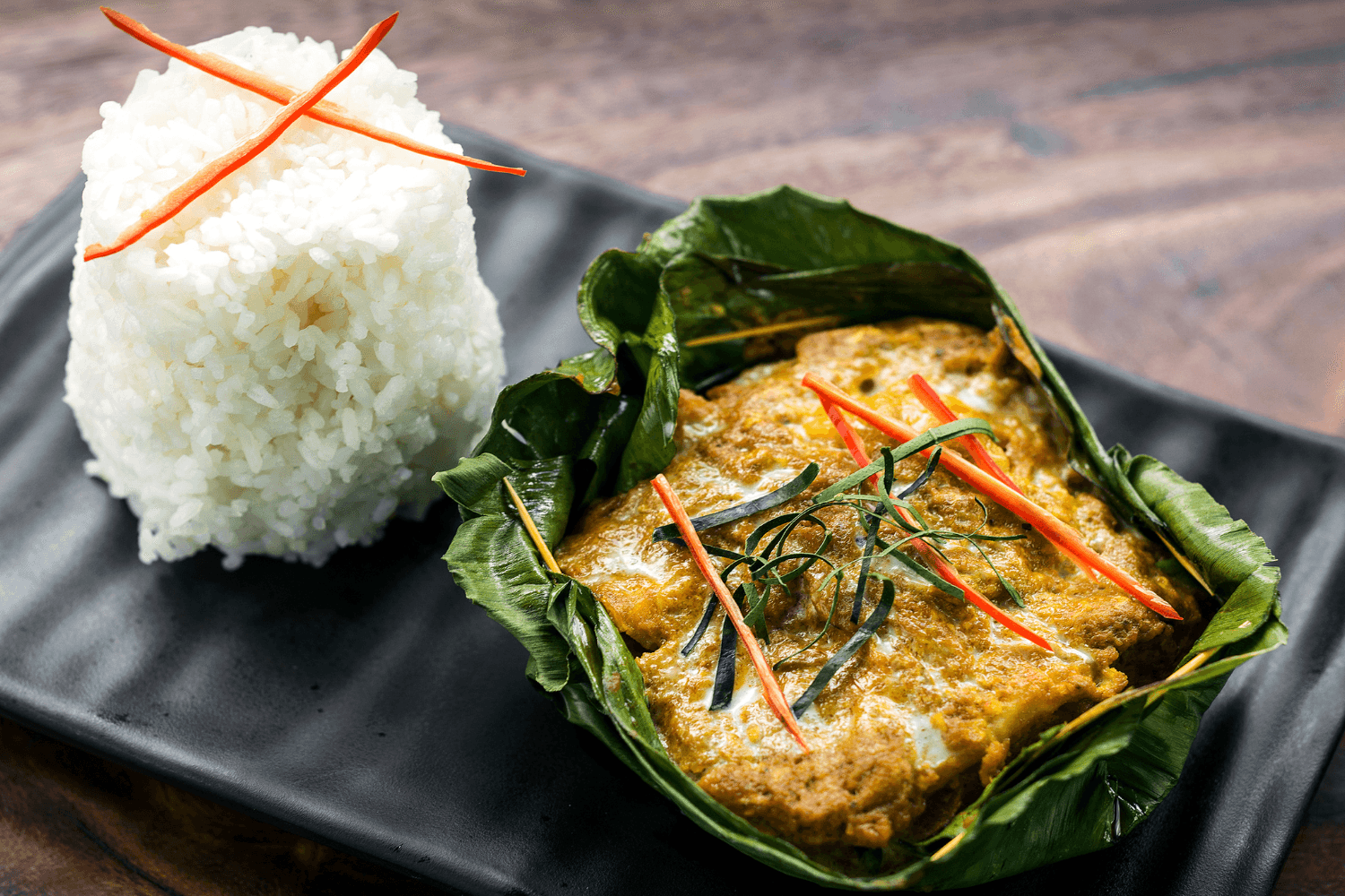 Cambodian fish amok steamed in banana leaf with herbs and chili, served with a mound of rice on a black plate.