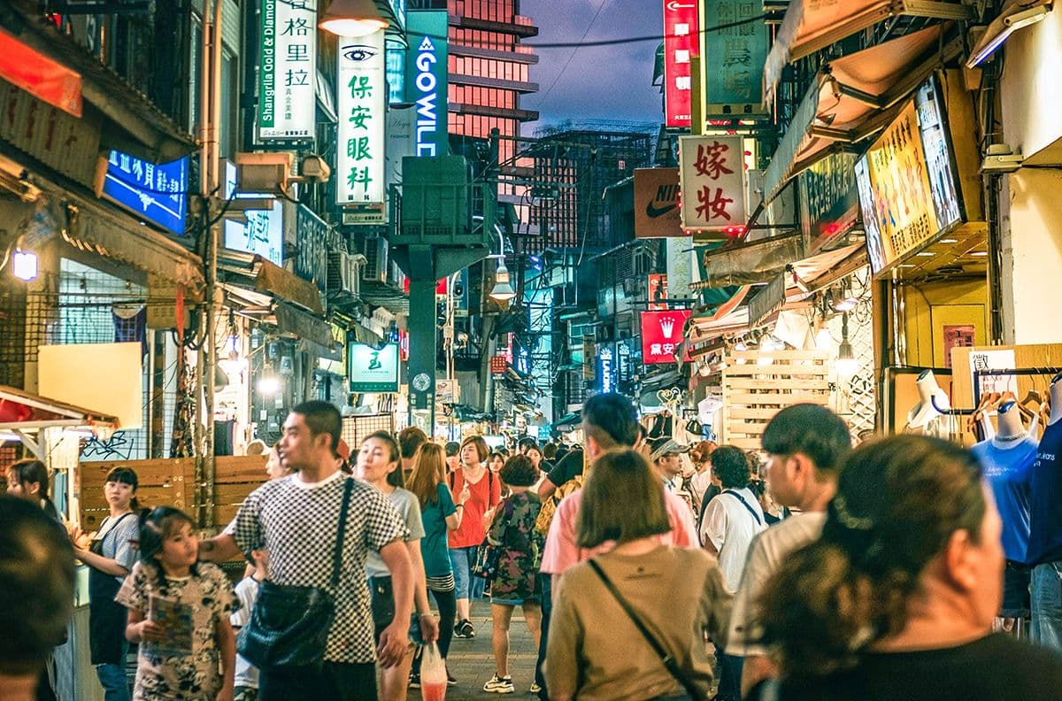 Crowded Shilin Night Market street in Taipei, Taiwan, lit by neon signs and vendor lights, with shoppers walking between food stalls and small shops.