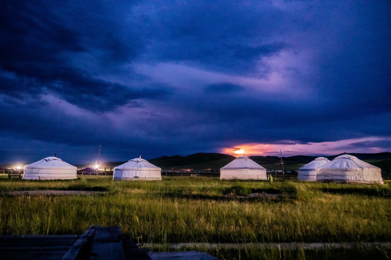 White Mongolian gers on open grassland at dusk under a dramatic blue sky, with sunset light on the horizon.